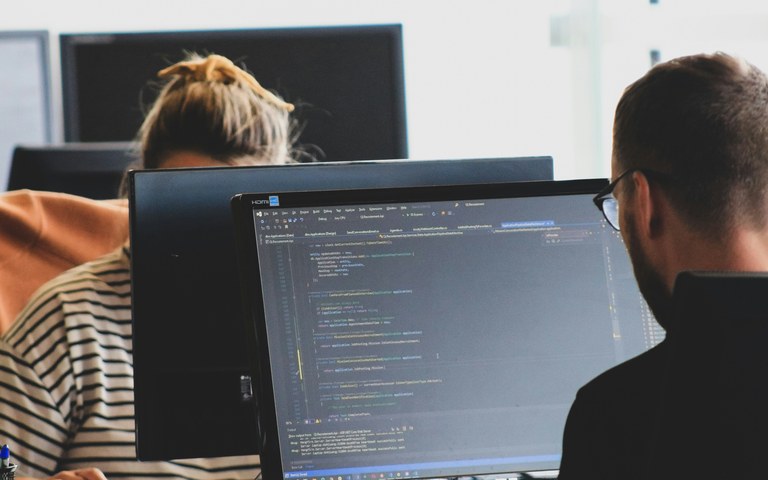 Two developers sit in a bright office facing several monitors displaying source code.
